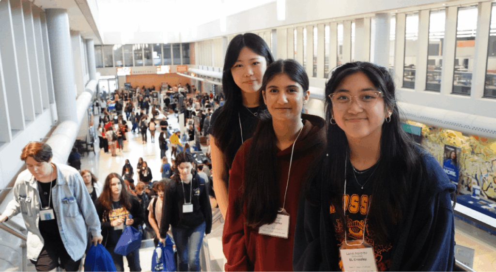 Three students standing on an escalator at a pathways fair, with attendees and information booths visible in the background.