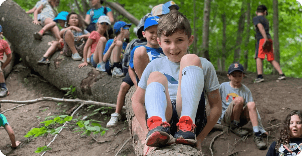 Children participating in an outdoor education activity, sitting and walking on a large fallen tree in a wooded area.